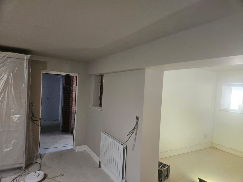 Interior painting in progress with light grey walls, white trim, a new radiator and furniture covered in protective plastic sheeting.