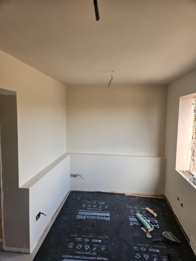 Small cream-painted room with professional black floor protection sheets, a paint roller and tools during an interior job.
