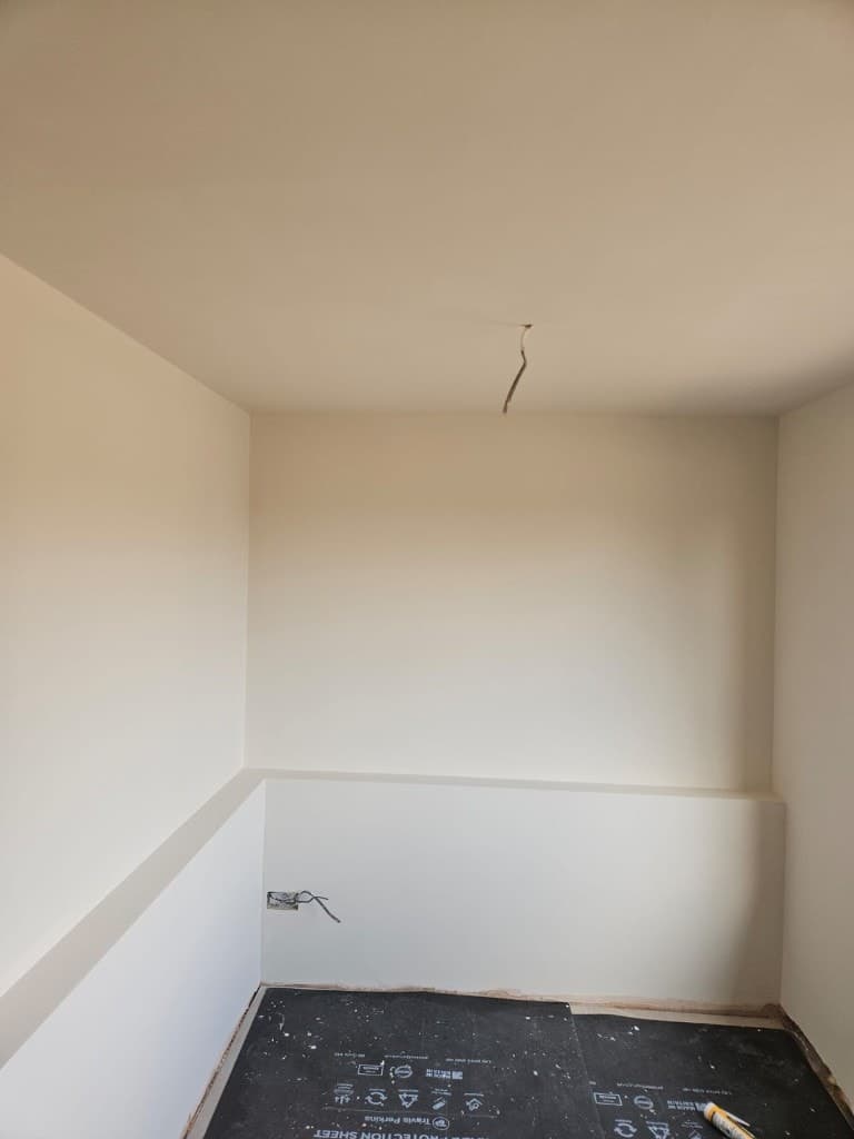 Small cream-painted room with a built-in wall ledge, exposed wiring and trade floor protection during renovation.