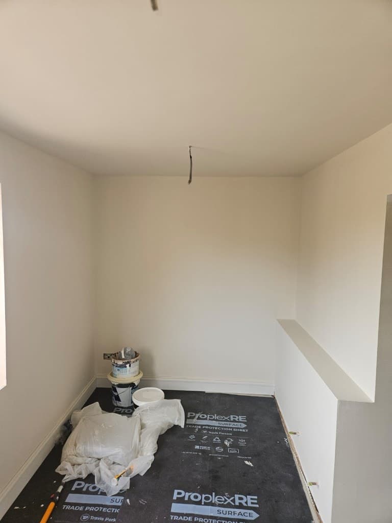 Small freshly painted cream alcove with white skirting, black floor protection, paint buckets and dust sheets.